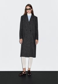 Šedý vlněný dvojřadý kabát s velkými kapsami, nošený přes světle modrý top a bílé legíny, doplněný tmavými sandály na podpatku se zavazováním.