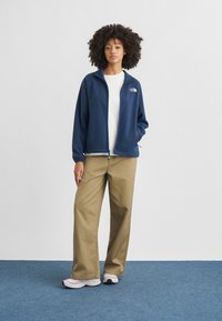 Ναυτικό μπουφάν από φλις με φερμουάρ, άσπρο T-shirt από κάτω, μπεζ φαρδιά παντελόνια και ανοιχτά αθλητικά παπούτσια, σε ουδέτερο φόντο.