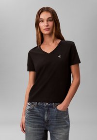 T-shirt nera in cotone con scollo a V e maniche corte, con un piccolo logo bianco "cK" sul petto a sinistra. Indossata con jeans di denim blu.