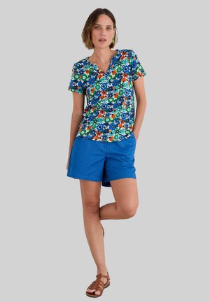 Top de manga corta con estampado floral sobre fondo azul, combinado con pantalones cortos azules. El top presenta un escote en V y un corte holgado. Sandalias marrones completan el look.