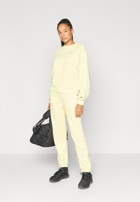 Κίτρινο φούτερ και αντίστοιχο παντελόνι φόρμας σε χαλαρή γραμμή, συνδυασμένα με μαύρα αθλητικά παπούτσια και μαύρη τσάντα duffel. Λεία υφή και απλός σχεδιασμός.