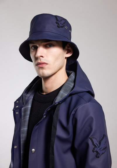 Marineblauer, wasserdichter Sonnenhut mit geprägt gestalteter Adler-Logo; kombiniert mit einer marineblauen Regenjacke mit karierter Futterkapuze und Druckknöpfen.