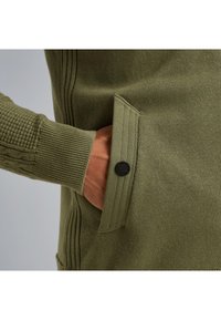 Olivno zeleni pulover z teksturiranim pletenjem, rebrastimi rokavi in stransko žepom z zapiranjem na črno gumb. Gladka tkanina.