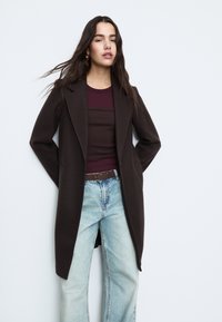 Tmavě hnědý kabát z vlněné směsi s přizpůsobeným střihem, s klopovým límcem, předními kapsami, nošený přes světle modré džíny s širokými nohavicemi.