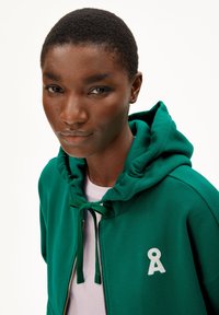 Grüne Zip-Hoodie mit verstellbarem Kapuzenzug, mit einem weißen Logo auf der Brust und einem weichen, strukturierten Stoff. Einfaches Unterhemd sichtbar.