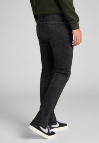 Man i slim-fit svarta jeans, en mörkgrön tröja och svarta sneakers med vita sulor och swoosh-logotyp, stående på en grå bakgrund.