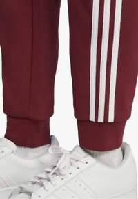 Vínové tepláky mají bílé třípruhy podél stran, v kombinaci s bílými sportovními botami. Tkanina vypadá měkce a má žebrované manžety.