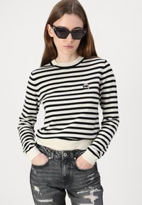 Black and white striped knit sweater with ribbed cuffs and hem, featuring a small embroidered logo. Worn with distressed black jeans.