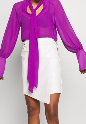 Donna che indossa una blusa trasparente viola a maniche lunghe con un foulard al collo, abbinata a una mini gonna bianca asimmetrica.