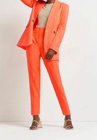 Neon orange skräddarsydd kavaj med dubbelknäppt design och matchande figurnära byxor. Slät textur, kombinerat med vita strappy klacksandaler.