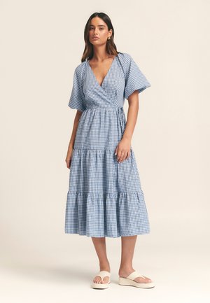 Zils un balts rūtainas raksta iesaiņojuma stila kleita ar īsām piedurknēm, kārtotu svārku un regulējamu jostas siksnu. Modelis valkā neitrālas krāsas sandales.