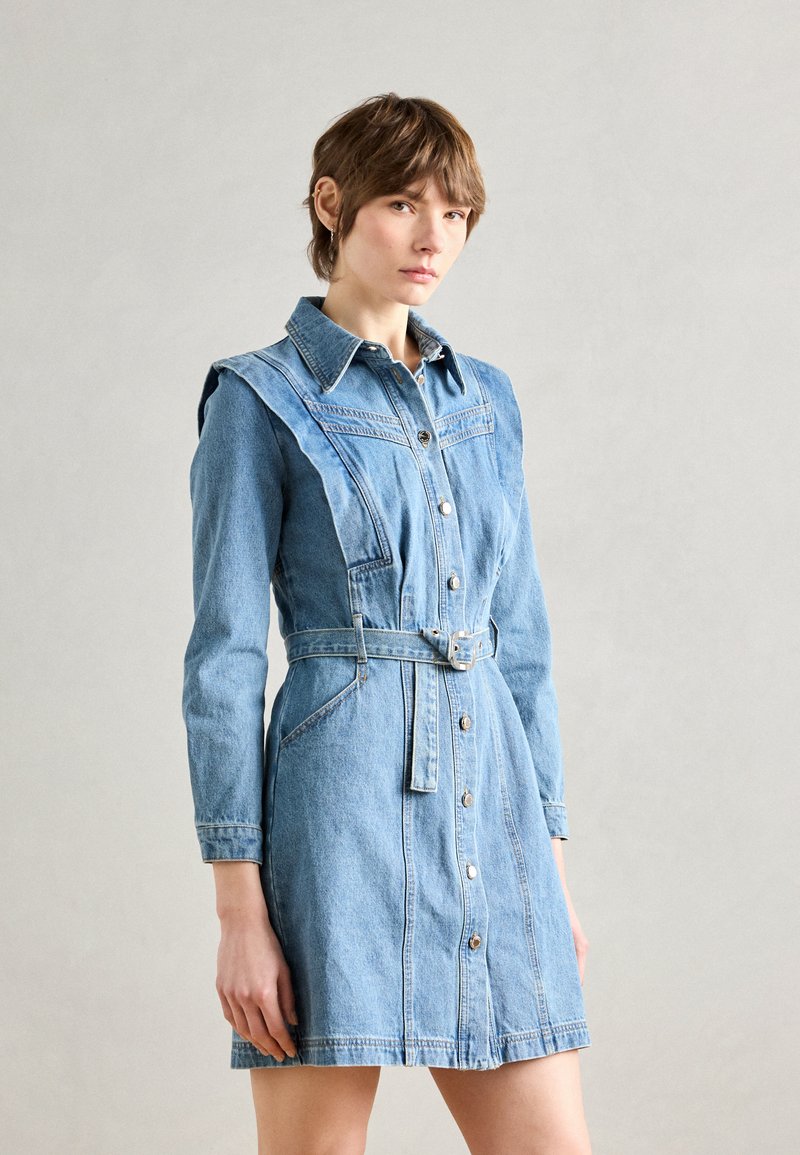 Femme portant une robe en jean avec ceinture, boutonnée sur le devant, manches longues et col pointu, debout sur un fond uni gris clair.