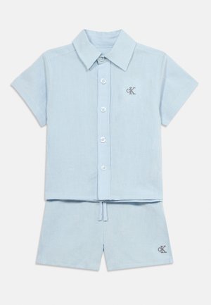 Svijetloplava košulja kratkih rukava na gumbe i odgovarajuće kratke hlače sa Calvin Klein "cK" logom na prsima košulje i nogavici kratkih hlača.