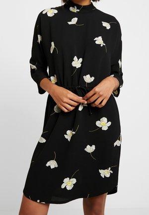 Frau trägt schwarzes Kleid mit weißem und gelbem Blumenmuster und Dreiviertelärmeln, bindet den Taillengürtel.
