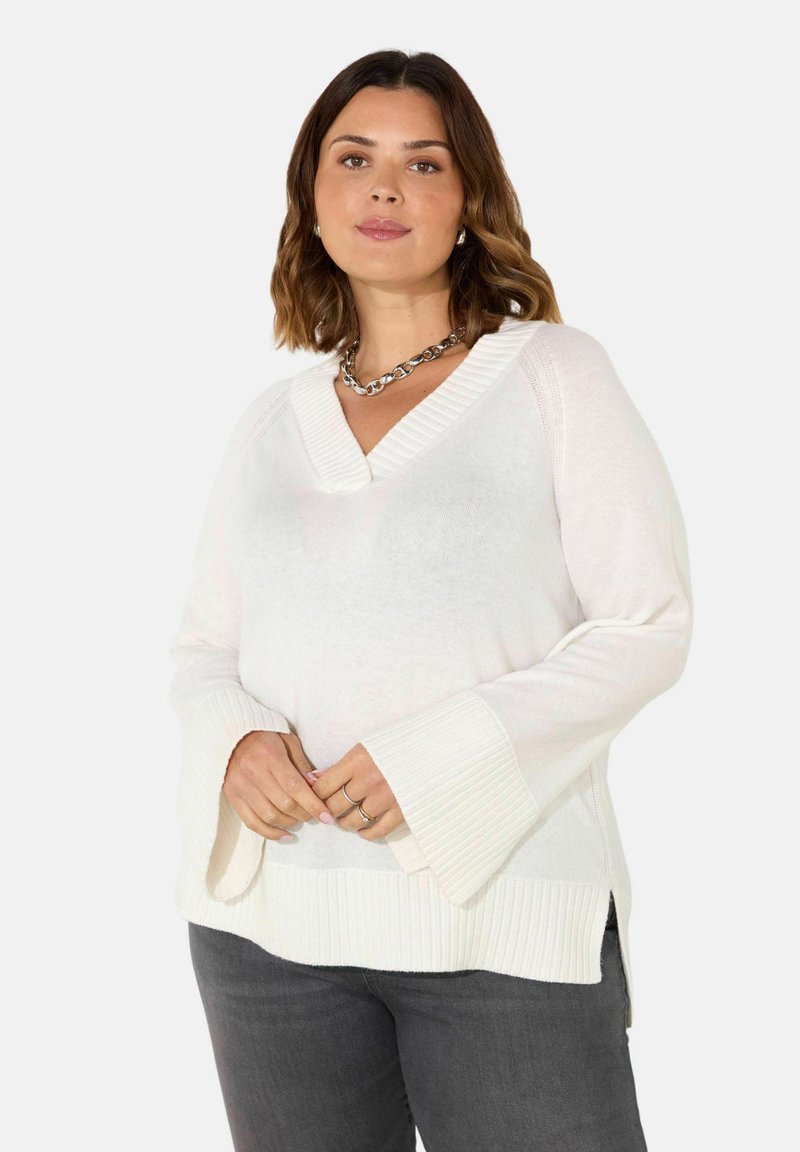 Maglione bianco lavorato a maglia con scollo a V, polsini a costine e spacchi laterali. Tessuto morbido, con maniche a campana. Abbinato a pantaloni grigi.