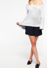 Biały sweter z dzianiny w oversize'owym stylu, z dekoltem w kształcie litery V i szerokimi rękawami, w zestawieniu z granatowymi szortami i czarnymi mokasynami.