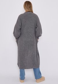 Langer, grauer Cardigan aus strukturiertem Strickstoff. Mit weiten Ärmeln, seitlichen Taschen und einer lässigen Passform, die unter die Knie reicht.
