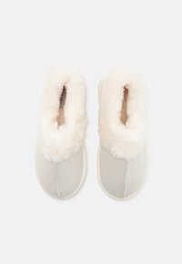 Lys grå hjemmesko med en fluffy hvid kunstlæderkant, blødt tekstiloverdel og kontrasterende stikninger langs sålens kant.
