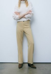 Pantalons beiges ajustés avec une taille haute et des plis avant, associés à un pull crème texturé. Des bottines à talons marron complètent le look.