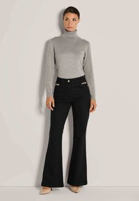 Frau in einem grauen Rollkragenpullover, schwarzen hoch taillierten Schlaghosen mit goldenen Kettendetails und beigen spitzen High Heels, die an einer Wand steht.