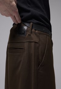 Pantalon en coton marron à coupe ajustée, avec un patch en cuir à l'arrière et des poches latérales, complété par une ceinture noire.