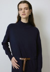 Kék kötött pulóver magas nyakkal, hosszú ujjal és barna övvel a deréknál. Bordás mandzsettával és sima textúrával rendelkezik.