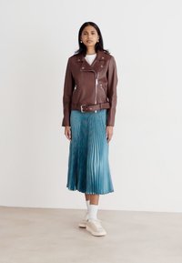 Jachetă de motociclist din piele maro, cu fermoare argintii, asortată cu o fustă plisată albastră. Accesoriile includ șosete albe și adidași de culoare deschisă.