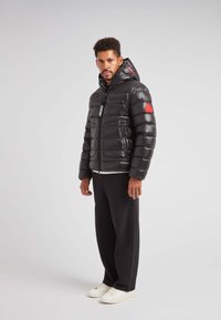 Giacca a vento nera con una texture lucida, chiusura con zip e dettaglio rosso sulla manica. Indossata sopra una camicia bianca, abbinata a pantaloni neri e sneakers bianche.