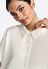 Blouse blanche à manches courtes avec col pointu, dotée d'une fermeture à boutons sur le devant et d'un tissu lisse. Finition légèrement brillante et coupe décontractée.