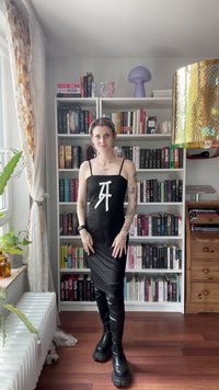 Juoda be rankovių suknelė su baltu grafiniu dizainu, blizgantys juodi iki kelių batai ir matomi tatuiruotės. Fone yra knygų lentynos.
