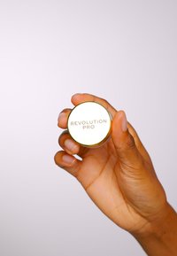 Rund gullcompact med en hvit topp merket "REVOLUTION PRO," holdt i en hånd. Compacten har en jevn overflate og blank finish.