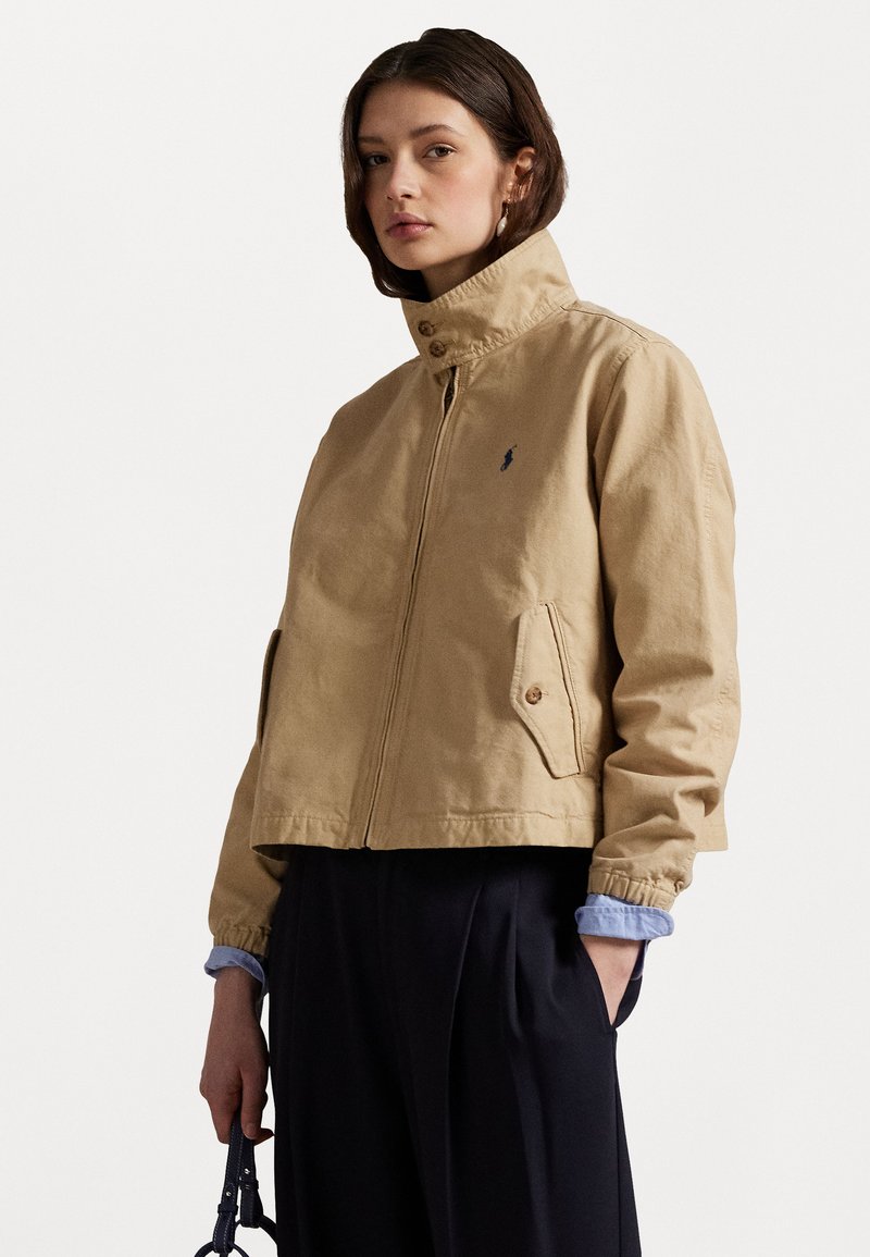 Beige kurz geschnittener Baumwolljacke mit Stehkragen, Reißverschluss, seitlichen Taschen und kleinem Logo auf der linken Brust. Strukturierten Oberfläche.