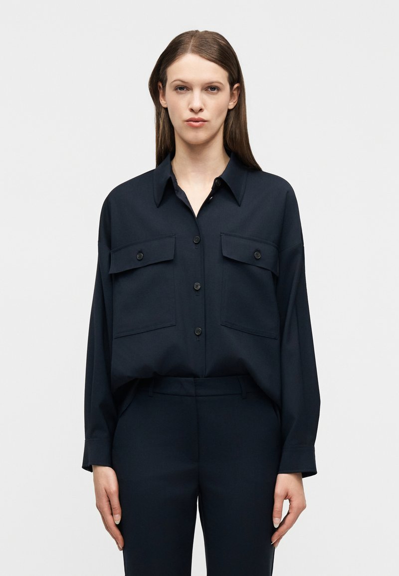 Γυναίκα με ίσια καστανά μαλλιά, που φοράει σκουρό μπλε πουκάμισο με κουμπιά και τσέπες με καπάκι, καθώς και παντελόνι που ταιριάζει, στέκεται μπροστά σε ένα σκέτο φόντο.