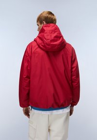 Ελαφρύ κόκκινο μπουφάν με κουκούλα και χαλαρή εφαρμογή, με ελαστικό τελείωμα και μανσέτες, φορεμένο πάνω από μπλε και λευκό πολυεπίπεδο τοπ και μπεζ παντελόνια.