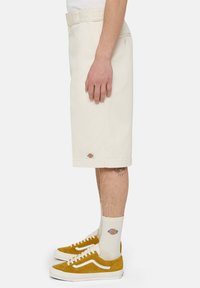 Shorts beiges en coton, avec une coupe droite et à longueur genou. Portés avec des baskets jaunes et des chaussettes blanches, mettant en avant un détail de logo.