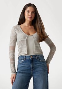 Cardigan tricotat de culoare gri deschis, cu mâneci lungi, cu modele geometrice și un design deschis în față, asortat cu blugi albastri cu talie înaltă.