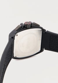 Πίσω μέρος μαύρου ρολογιού χειρός Hugo που δείχνει μεταλλική πίσω πλευρά με βίδες, λουράκι από υφασμένο καουτσούκ και κορώνα με κόκκινο δαχτυλίδι με την επιγραφή "Hugo".