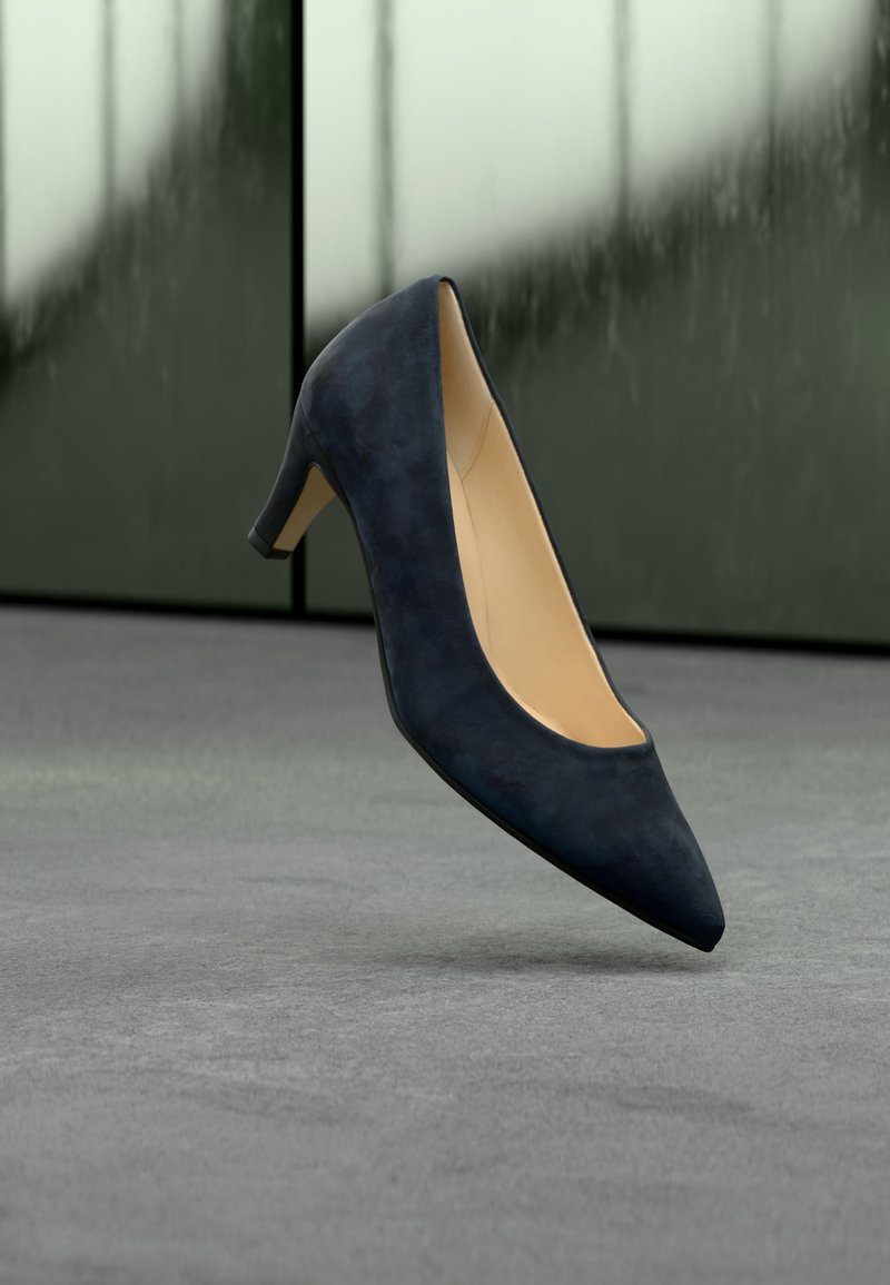 Chaussure femme en daim bleu marine à bout pointu et talon moyen flottant au-dessus d’un sol gris avec un fond vert-gris flou.