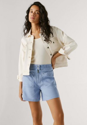 Pikkade tumedate juustega naine, kes kannab valget lühikest jopet, valget topi ja helesiniseid kõrge vöökohaga lühikesi pükse, poseerides neutraalse tausta ees.