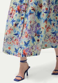 Květinové maxi šaty s pastelově zeleným základem, zdobené modrými, červenými a fialovými květy. Obsahují hnědé knoflíky a plisované detaily. Spárovány s modrými sandály na podpatku.