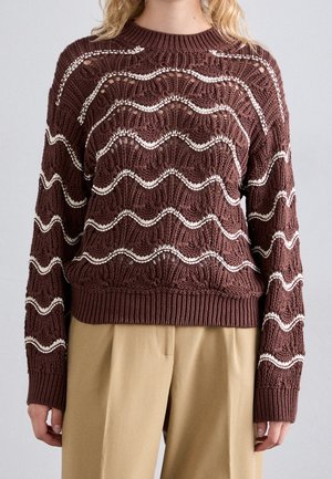 Pull en maille marron avec des rayures blanches ondulées et des détails ajourés texturés, porté avec un pantalon beige.