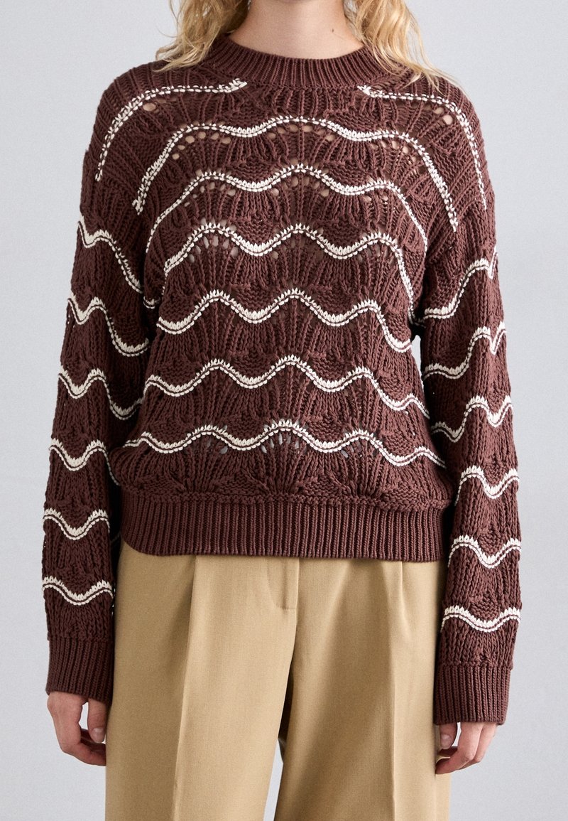 Pull en maille marron avec des rayures blanches ondulées et des détails ajourés texturés, porté avec un pantalon beige.