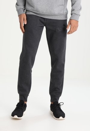 Mann trägt dunkelgraue Jogginghosen, graues Sweatshirt und schwarze Sneakers und steht vor einem schlichten weißen Hintergrund.