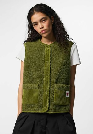 Jeune femme aux cheveux bouclés et foncés porte une veste en polaire vert olive boutonnée sur une chemise blanche à manches courtes, les mains dans les poches d'un pantalon noir.