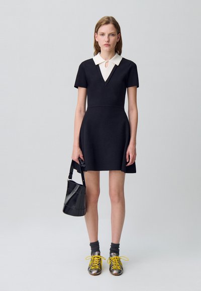 Vestito A-line nero con maniche corte e colletto bianco, caratterizzato da una scollatura a V e un dettaglio a chiave. Abbinato a scarpe metalliche e una borsa nera.