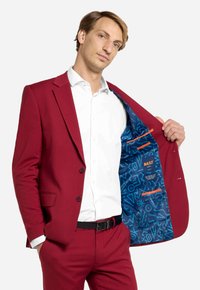 Giacca di tweed bordeaux con una fodera a fantasia azzurra chiara, dotata di due bottoni e tasche laterali. Camicia bianca sotto con una cintura nera.