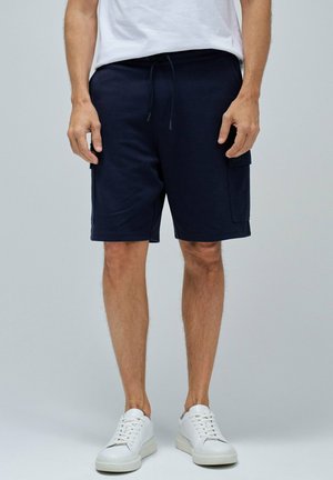 Shorts en coton bleu marine avec une taille élastique à cordon et des poches latérales, associés à des baskets blanches, présentés sur un fond gris.