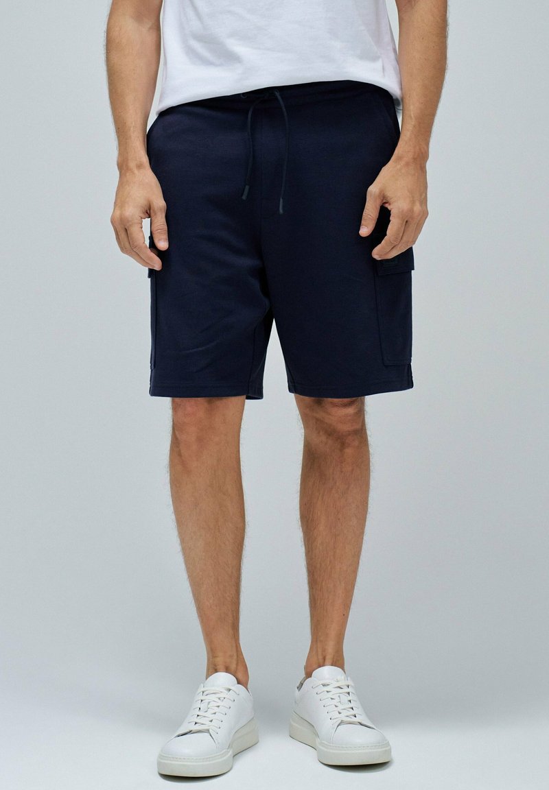 Pantalones cortos de algodón azul marino con cinturilla elástica y cordón ajustable, y bolsillos laterales, combinados con zapatillas blancas, presentados sobre un fondo gris.