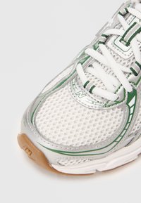 Gros plan d'une chaussure de sport blanche et verte avec tige en maille, lacets blancs et semelle en caoutchouc beige sur un fond blanc.