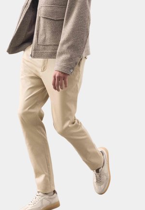 Beige Baumwollhosen kombiniert mit einer strukturierten, hellbraunen Jacke. Das Outfit wird durch cremefarbene Sneakers mit Gummisohle vervollständigt.